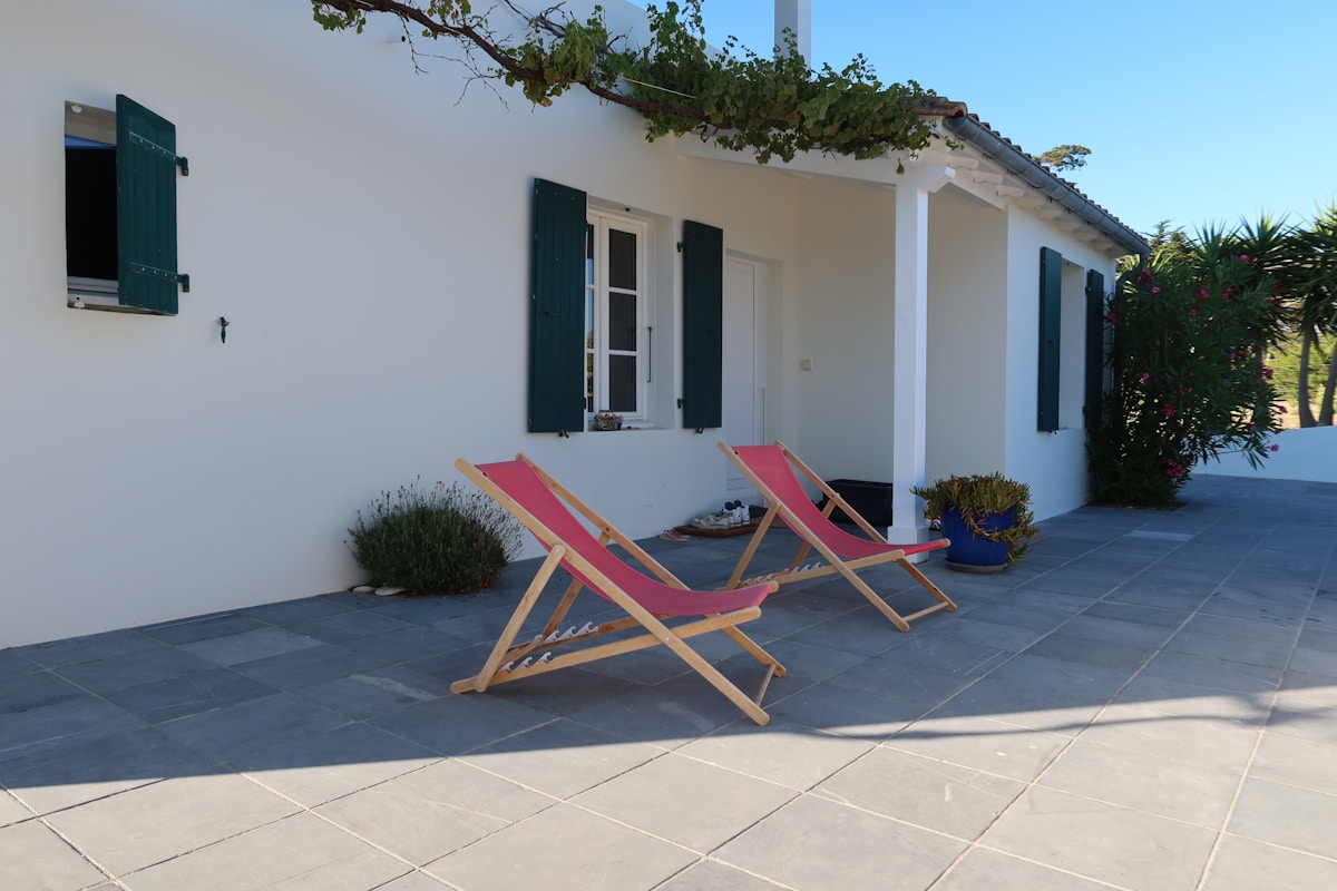 Maison blanche avec volets verts, chaises longues roses, jardin verdoyant.