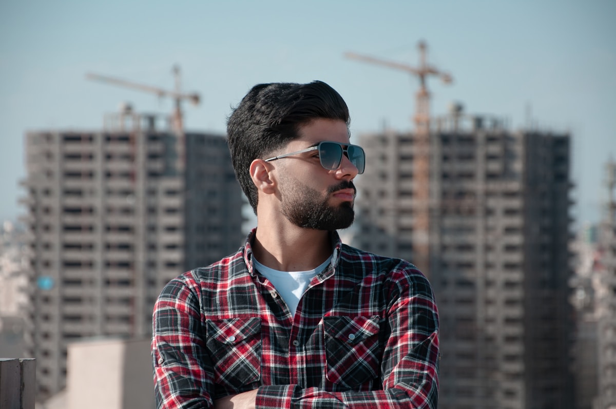 Homme avec barbe portant des lunettes de soleil sur un fond urbain.