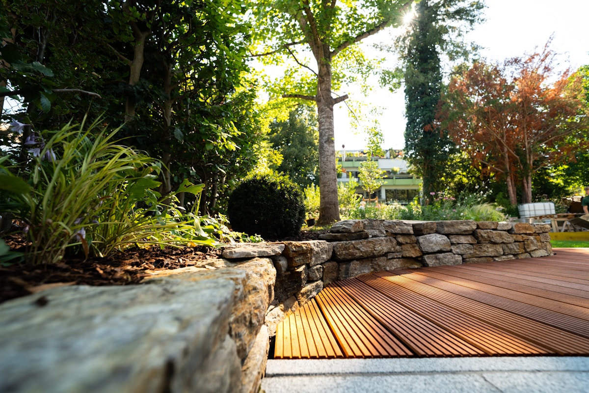 Jardin luxuriant avec terrasse en bois, murs de pierre et végétation verdoyante.
