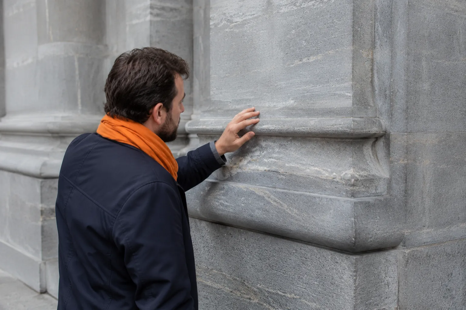 Homme en manteau bleu et écharpe orange touchant un mur en pierre.