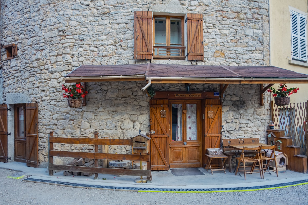 Maison de pierre avec fenêtres en bois, terrasse couverte et décoration florale.